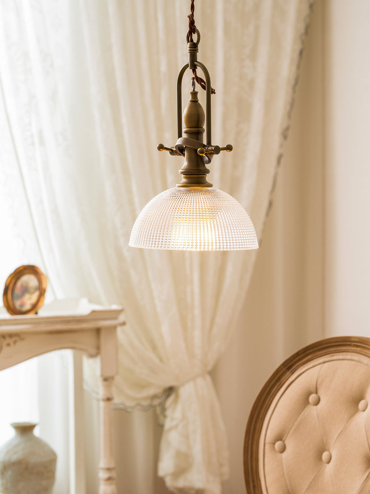 Lit Clérachic pendant beside a lace curtain, with a brass-framed mirror on a console and a cushioned chair at right.