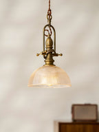 Clérachic pendant glowing amber on a cream wall, with its brass cradle, ribbed dome and a radio on a cabinet below.