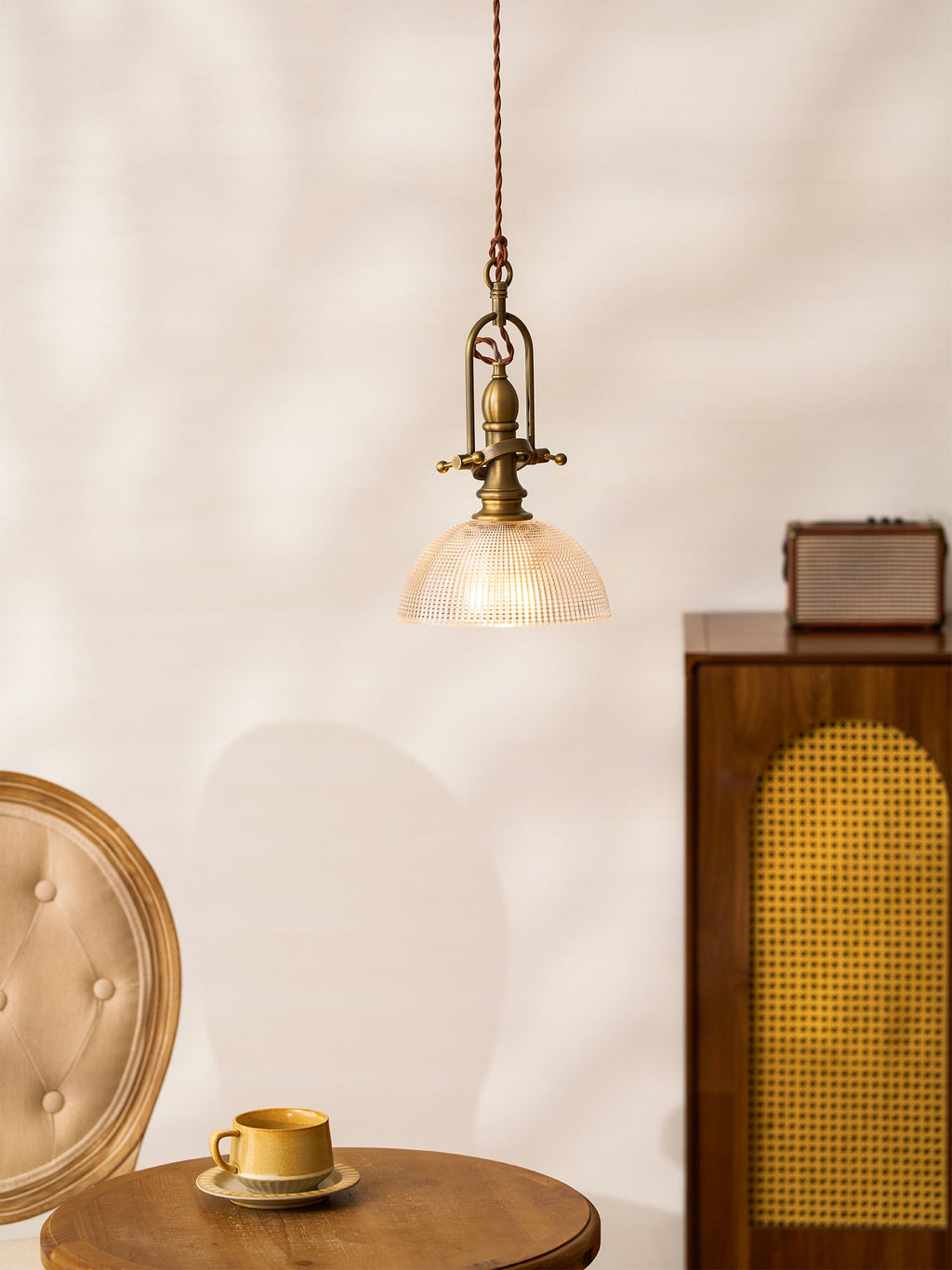 A small Clérachic pendant above a round table with a mustard cup and a cane-front cabinet, on a pale wall.