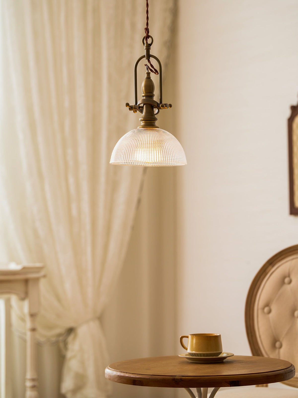 This Clérachic pendant hangs beside a lace curtain, with a button-back chair and a round wooden table with a cup below.