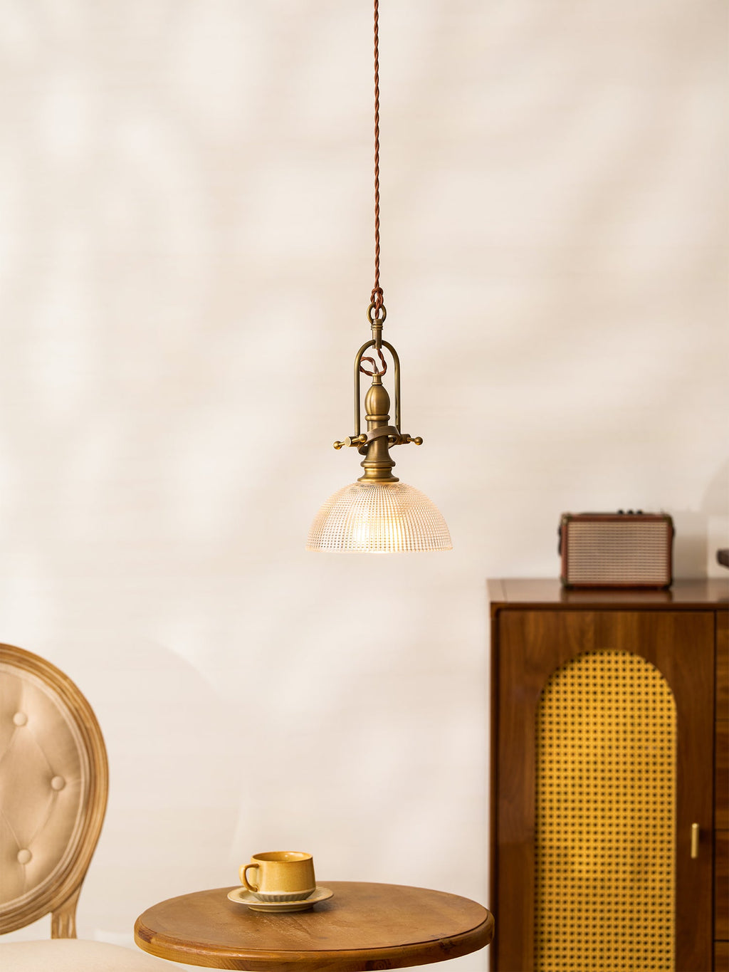 A Clérachic pendant with a clear ribbed dome, lit, next to a cane-front cabinet, a small radio and a round table.