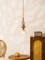 A Clérachic pendant with a clear ribbed dome, lit, next to a cane-front cabinet, a small radio and a round table.