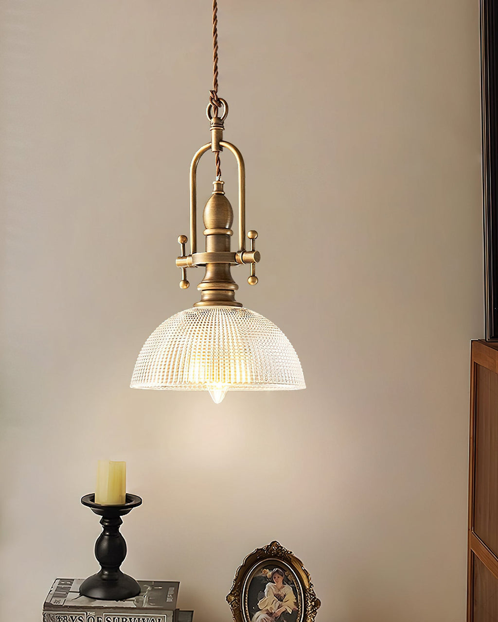 A Clérachic pendant hangs by a cream wall, with a candle on a black holder and an oval portrait frame below it.