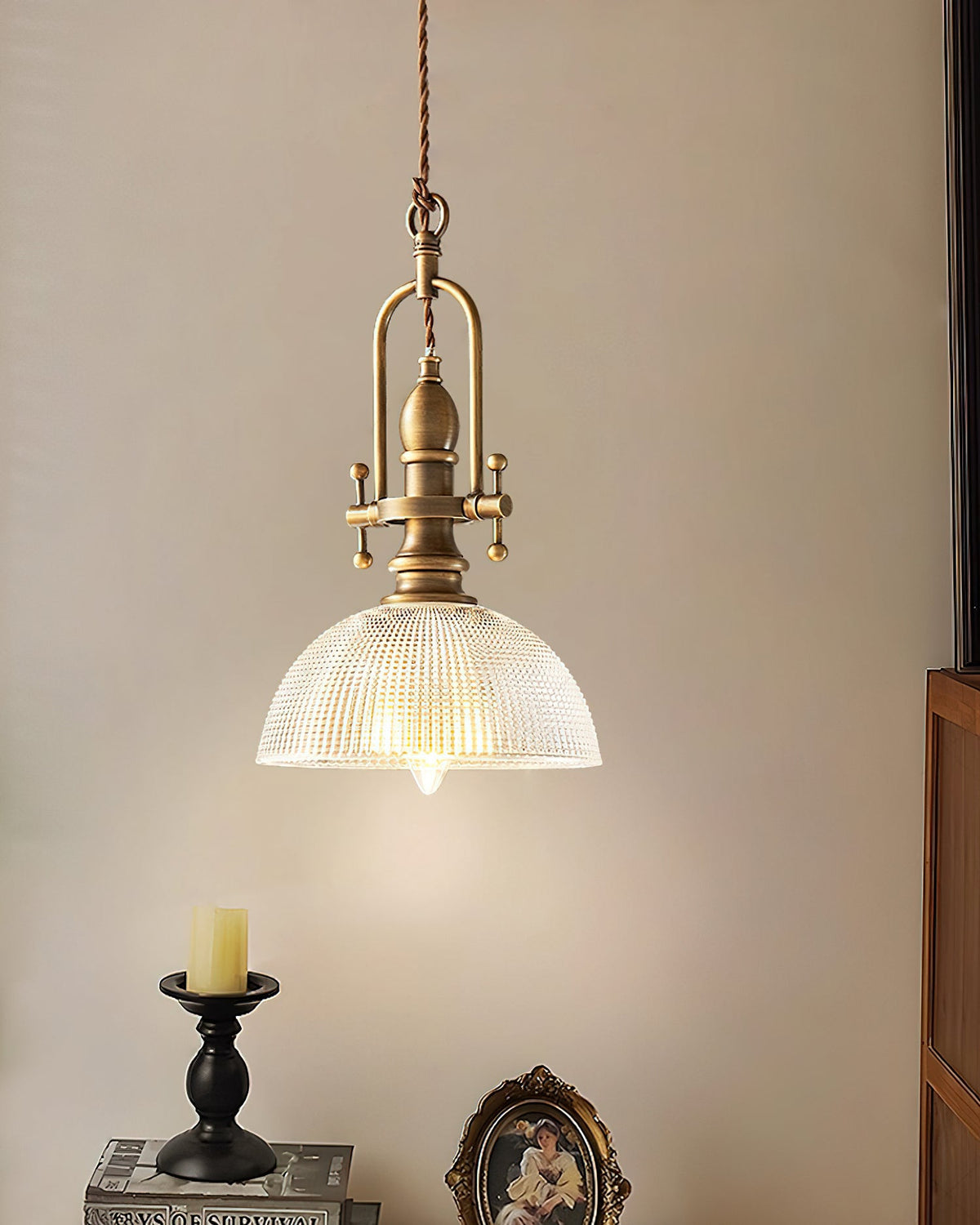 A Clérachic pendant hangs by a cream wall, with a candle on a black holder and an oval portrait frame below it.