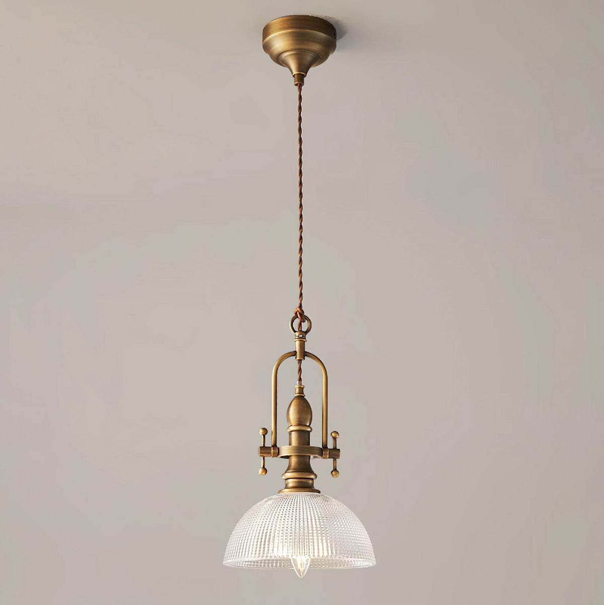 Clérachic pendant on a plain beige wall, with an antique brass canopy, a twisted brown cord and a clear ribbed dome.