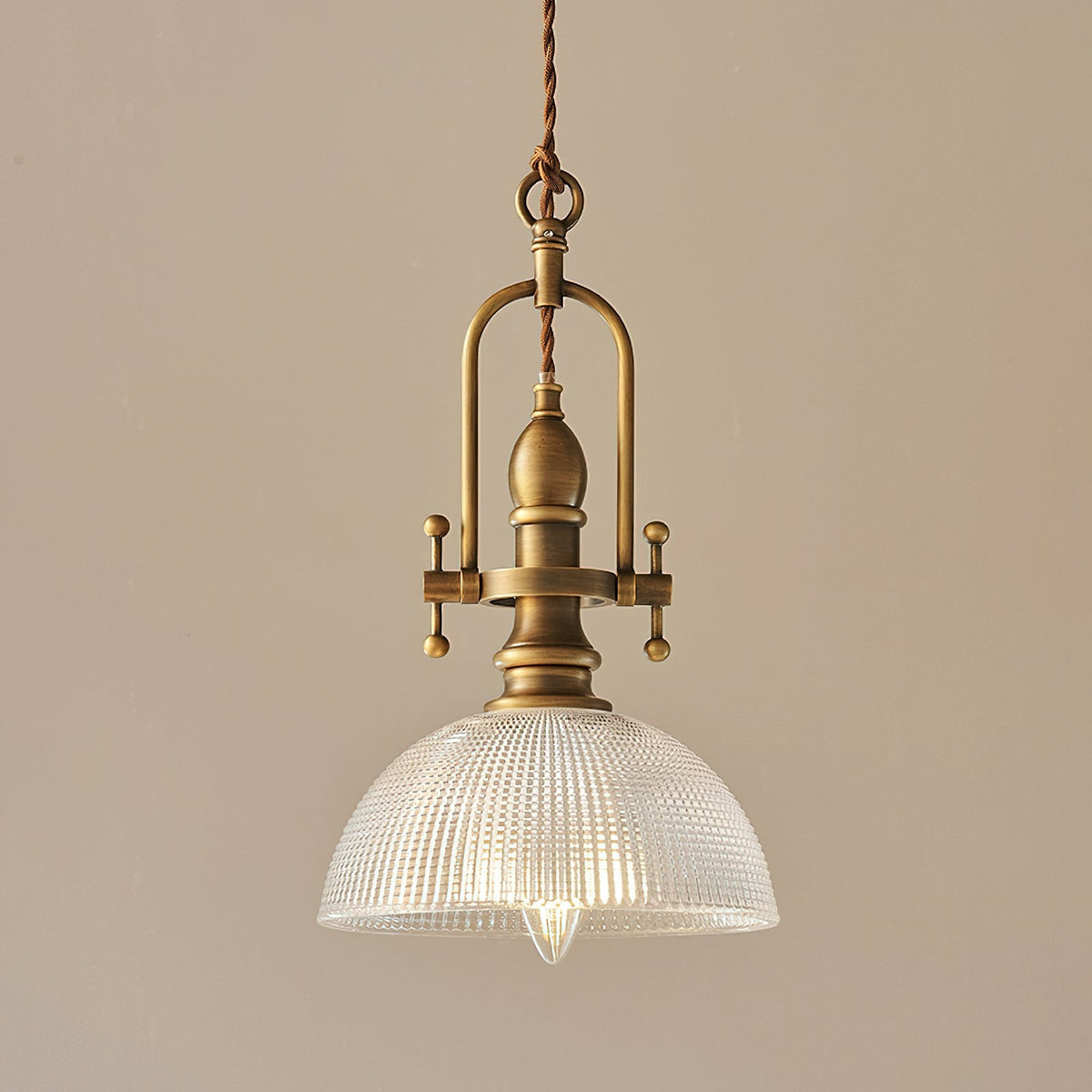 Clérachic pendant on a warm tan backdrop, with its brass frame and a ribbed glass dome lit around a filament bulb.