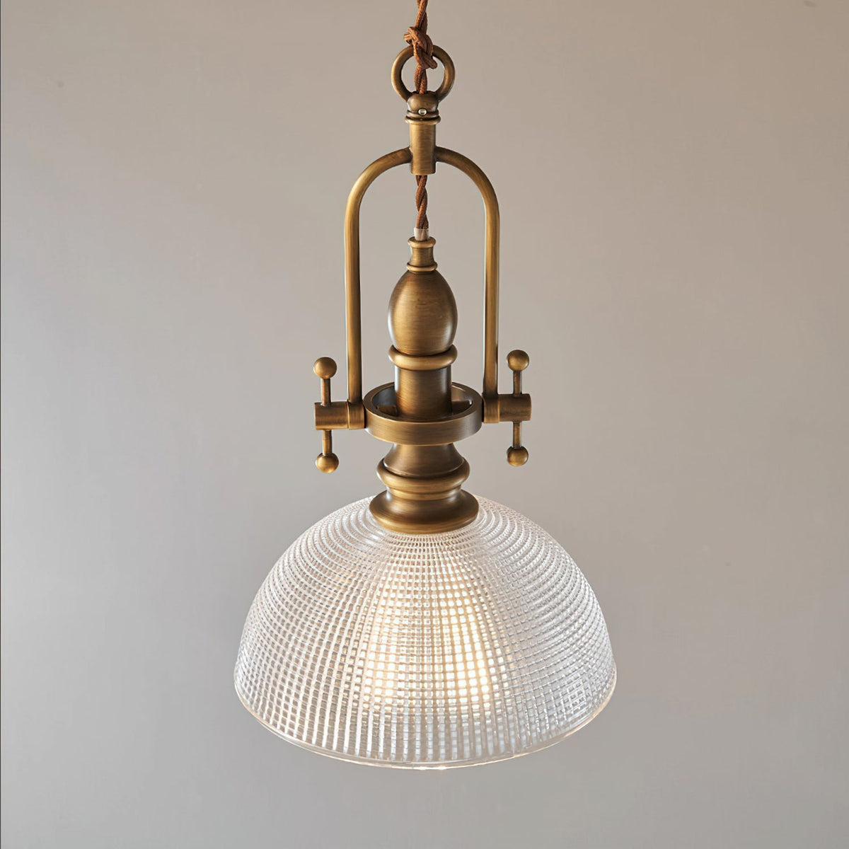 Close view of this Clérachic pendant's ribbed glass dome and brass cradle, lit, on a soft grey backdrop.