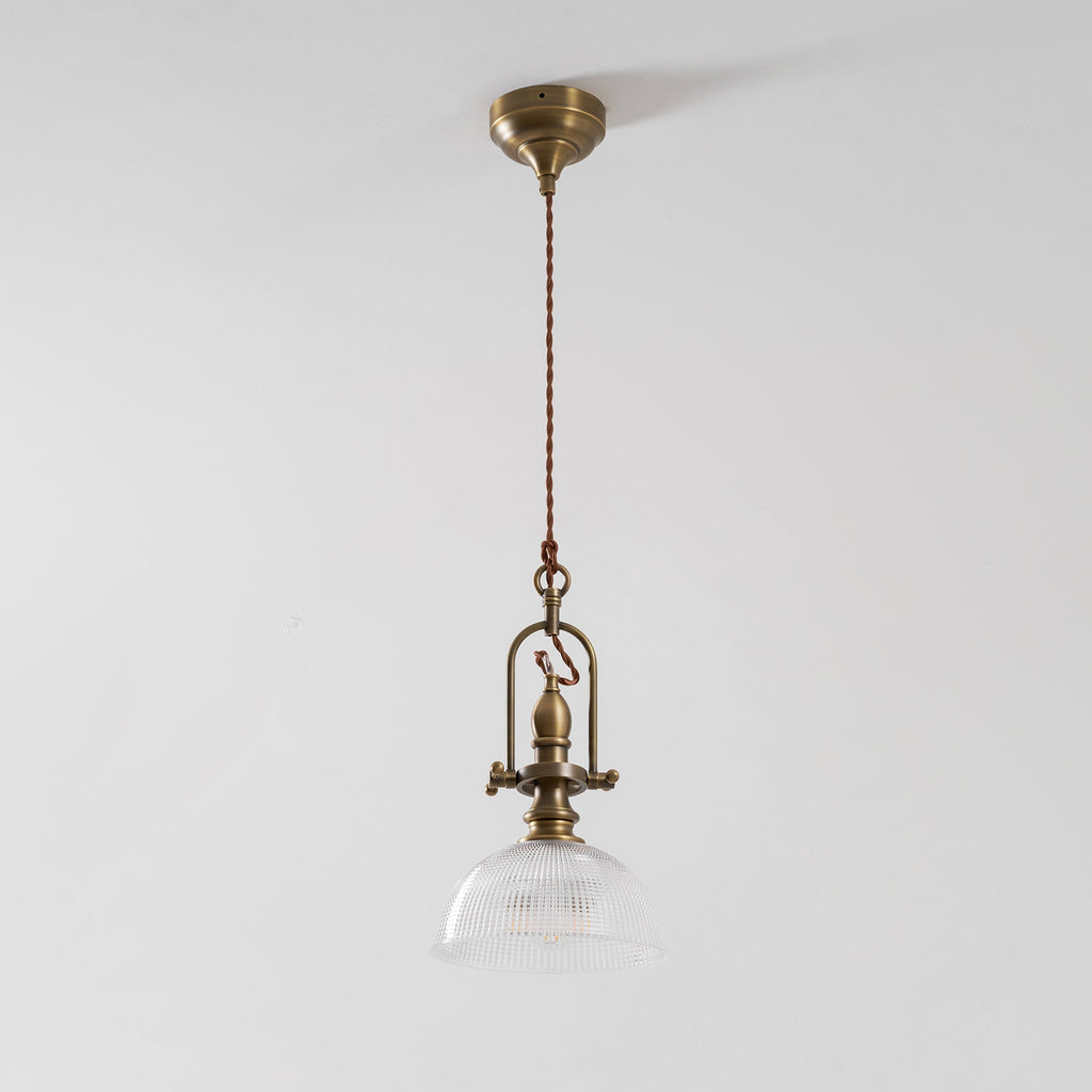 A Clérachic pendant with a brass canopy and long twisted cord, with the glass dome lit softly against a pale ceiling.