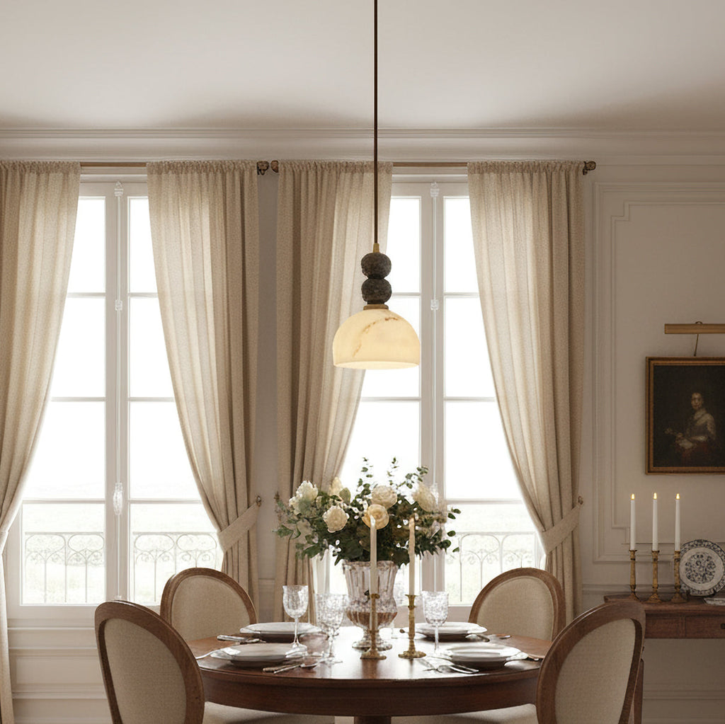 This Clérachic pendant light glows above a round dining table set with glasses and white roses, in front of tall linen curtains.