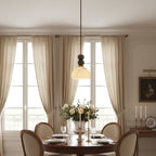 This Clérachic pendant light glows above a round dining table set with glasses and white roses, in front of tall linen curtains.