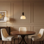 A lit Clérachic pendant light hangs over a round walnut table in a room with taupe panelled walls and cream dining chairs.