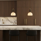 Two Clérachic pendant lights lit above a marble-topped kitchen island, in front of dark walnut cabinets with gold handles.