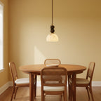 This Clérachic pendant light hangs low over a round wooden table with cane-backed chairs, against a warm yellow wall.