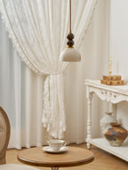 Unlit Clérachic pendant light with a pale dome shade, hanging in front of a lace curtain above a small round table with a teacup.