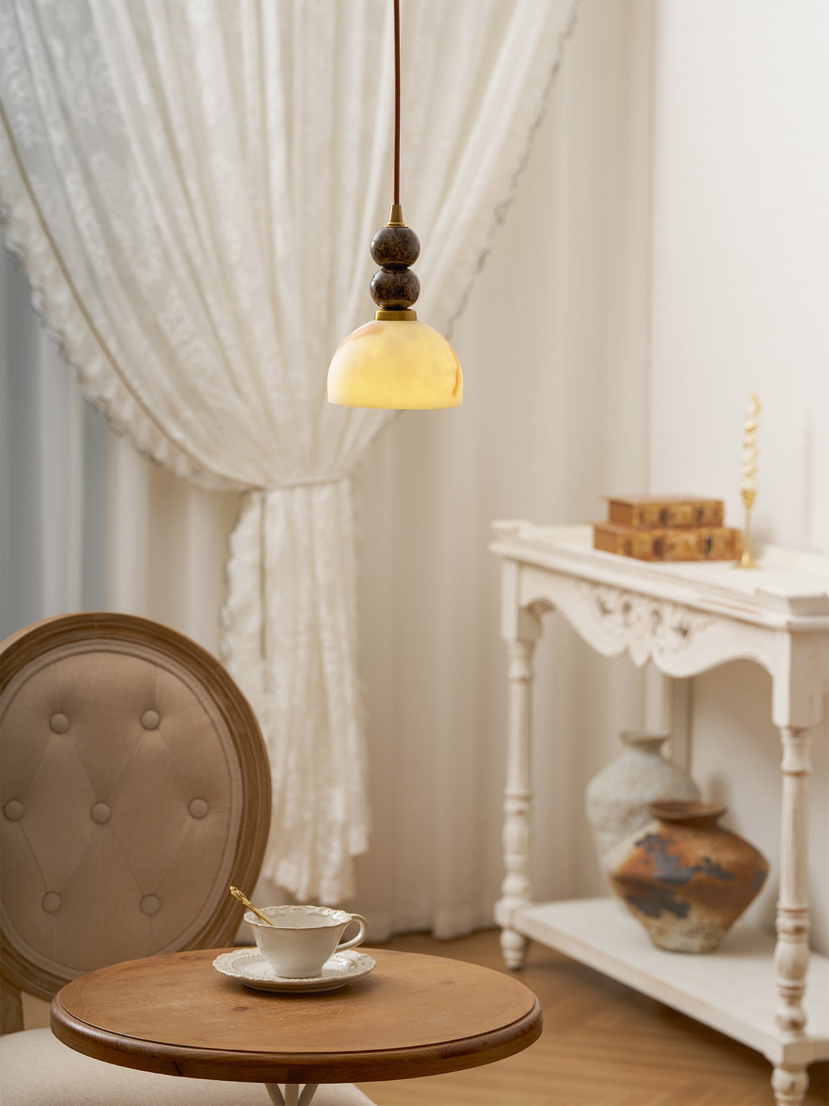 A Clérachic pendant light glowing yellow above a small round table with a teacup, beside a white carved console.