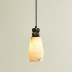 A Clérachic pendant light lit with a tapered, faceted shade glowing warm, below two small dark marble discs on a black cord.