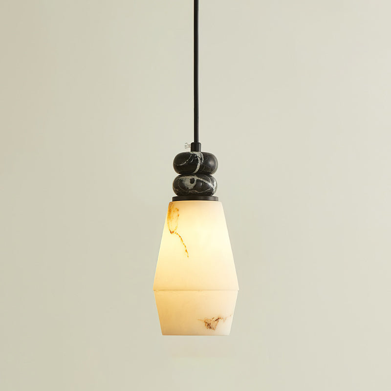A Clérachic pendant light lit with a tapered, faceted shade glowing warm, below two small dark marble discs on a black cord.