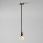 Full length of this Clérachic pendant light, lit, hanging on a thin brown cord from a brass ceiling plate against a grey wall.