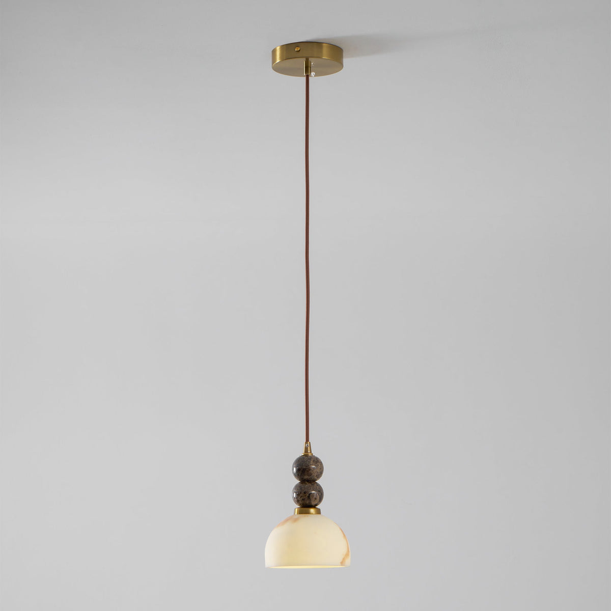 Full length of this Clérachic pendant light, lit, hanging on a thin brown cord from a brass ceiling plate against a grey wall.