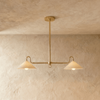 Clérachic pendant light in polished brass, its twin cone shades glowing warm against a plaster backdrop.