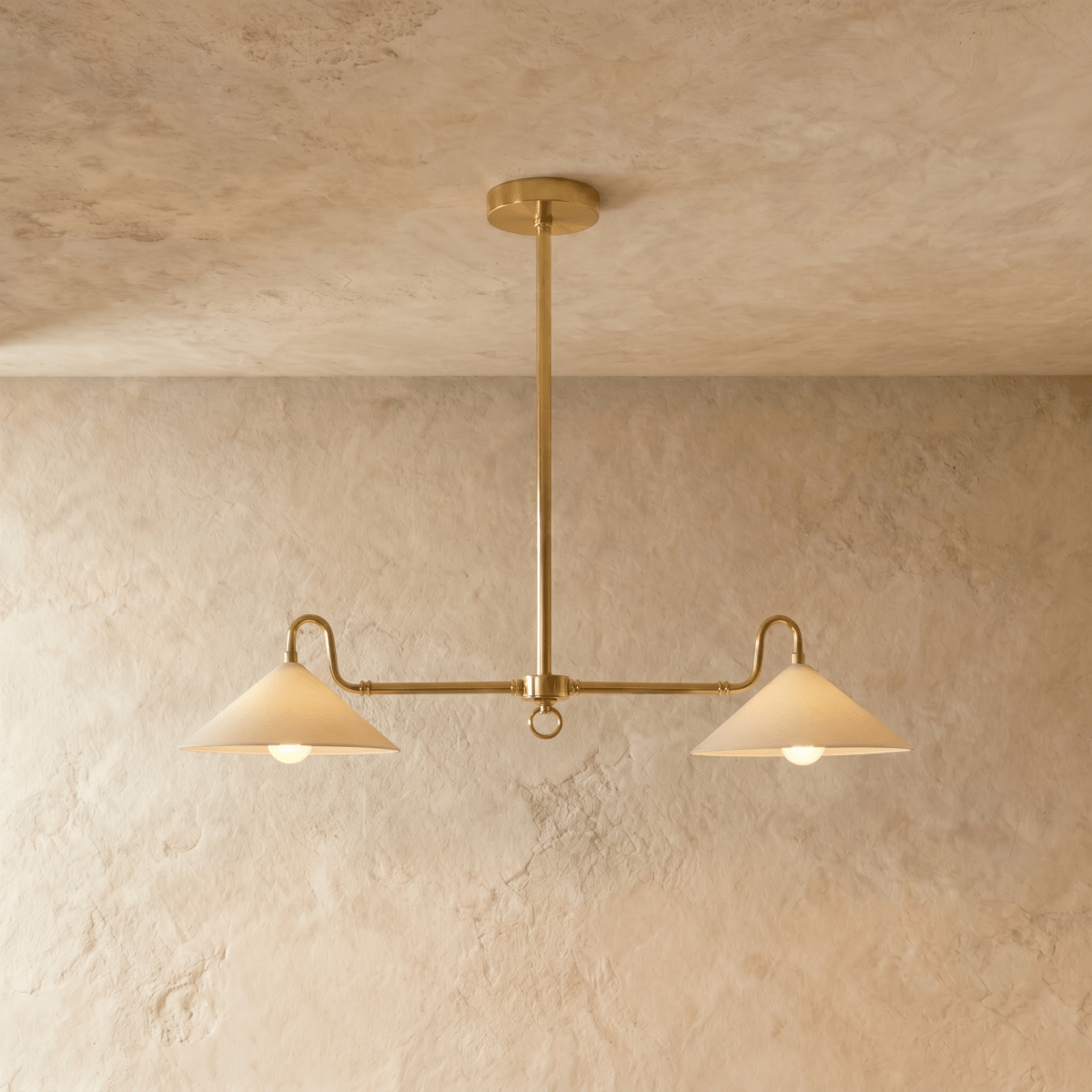Clérachic pendant light in polished brass, its twin cone shades glowing warm against a plaster backdrop.