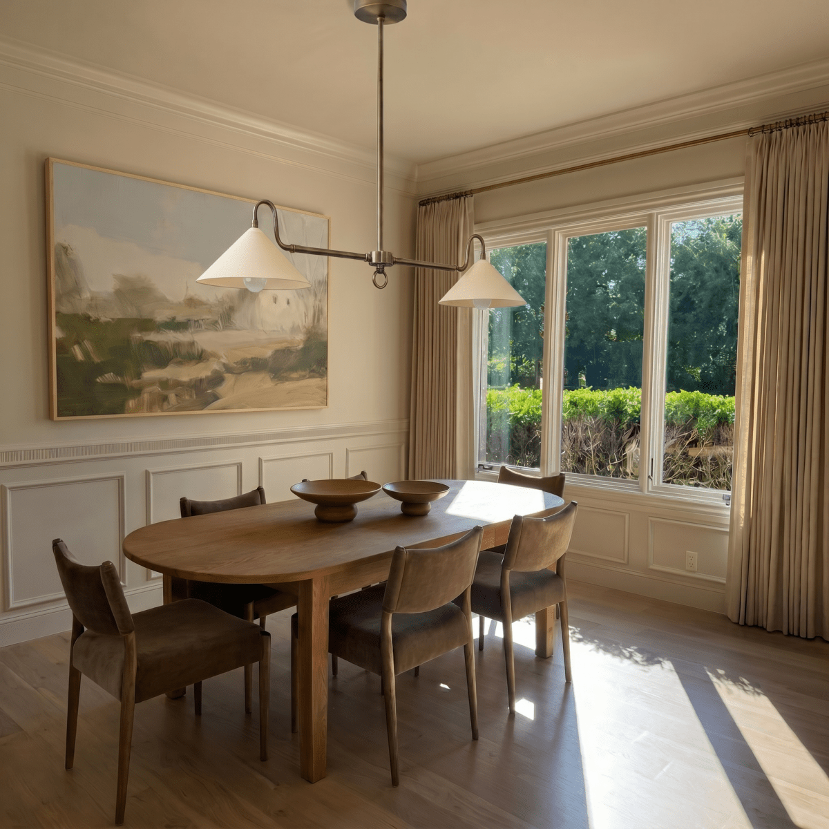 This chrome Clérachic pendant light hangs above an oval dining table beneath a framed landscape painting.