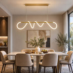 A Clérachic linear pendant with a lit silicone wave on a brass bar, centred above a marble dining table and a vase of greenery.