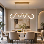 Brass bar pendant from Clérachic with a looping silicone ribbon lit white, above a dining table with taupe chairs and an arched alcove.
