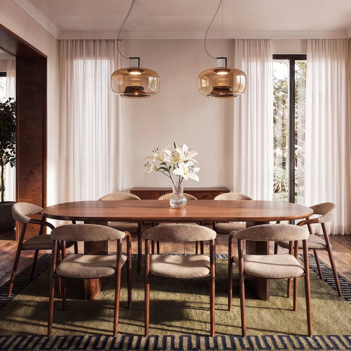 Two Clérachic amber glass pendants hung above a long walnut dining table with white lilies and sheer curtains.