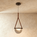 Clérachic pendant light with a tan leather strap holding a half-round smoked glass shade, hung from a dark ceiling canopy.