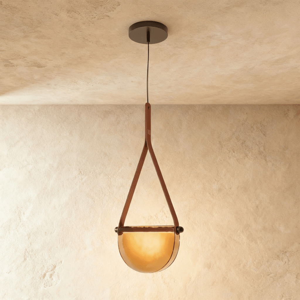 Clérachic pendant light with a tan leather strap holding a half-round amber glass shade that glows warm on a plaster ceiling.