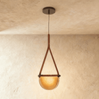 Clérachic pendant light with a tan leather strap holding a half-round amber glass shade that glows warm on a plaster ceiling.
