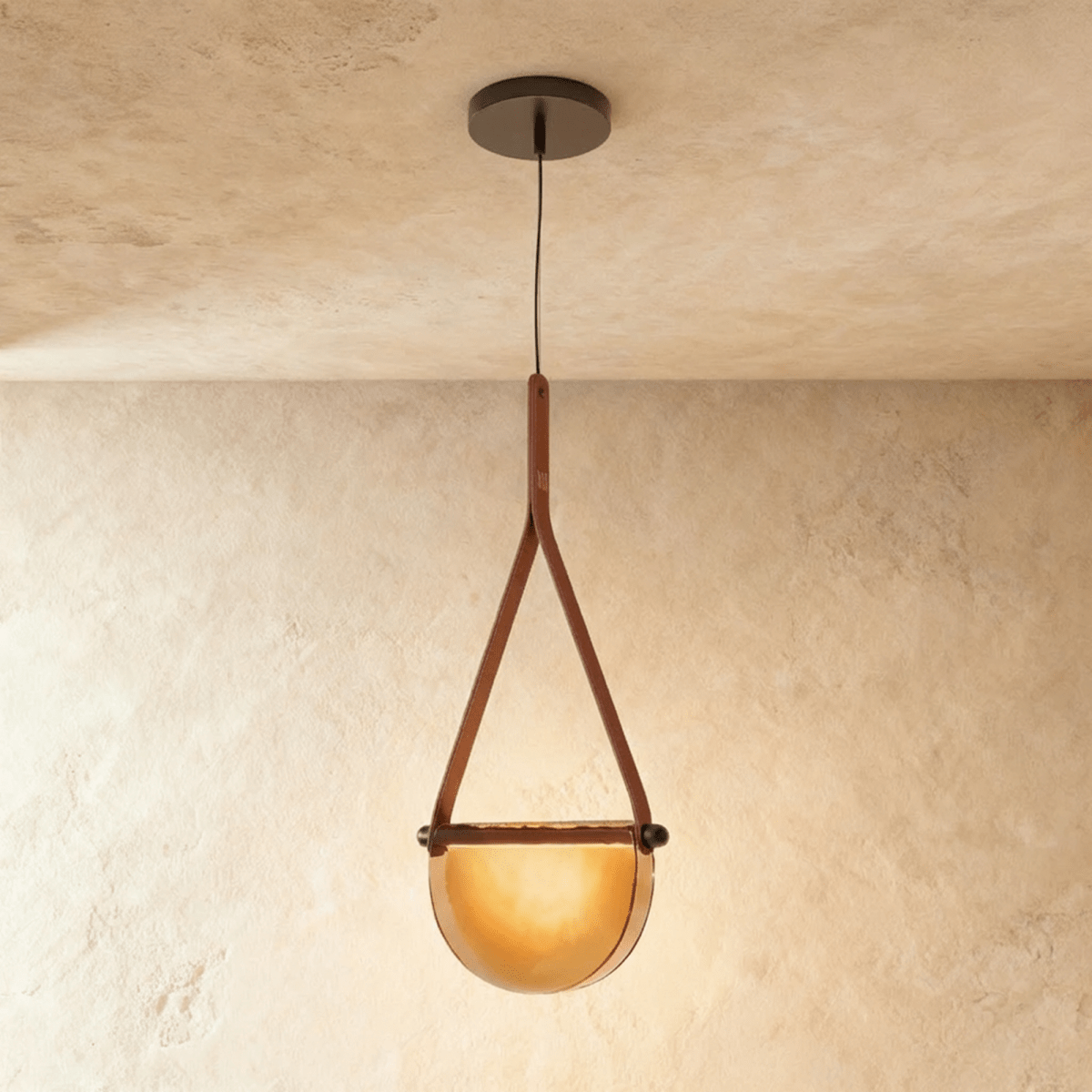 Clérachic pendant light with a tan leather strap holding a half-round amber glass shade that glows warm on a plaster ceiling.