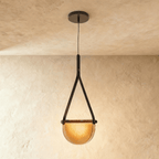 Clérachic pendant light with a black leather strap holding a half-round amber glass shade glowing warm on a plaster ceiling.
