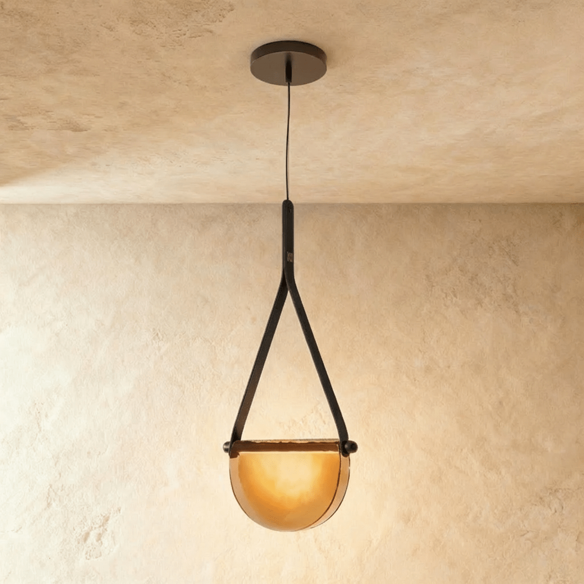 Clérachic pendant light with a black leather strap holding a half-round amber glass shade glowing warm on a plaster ceiling.