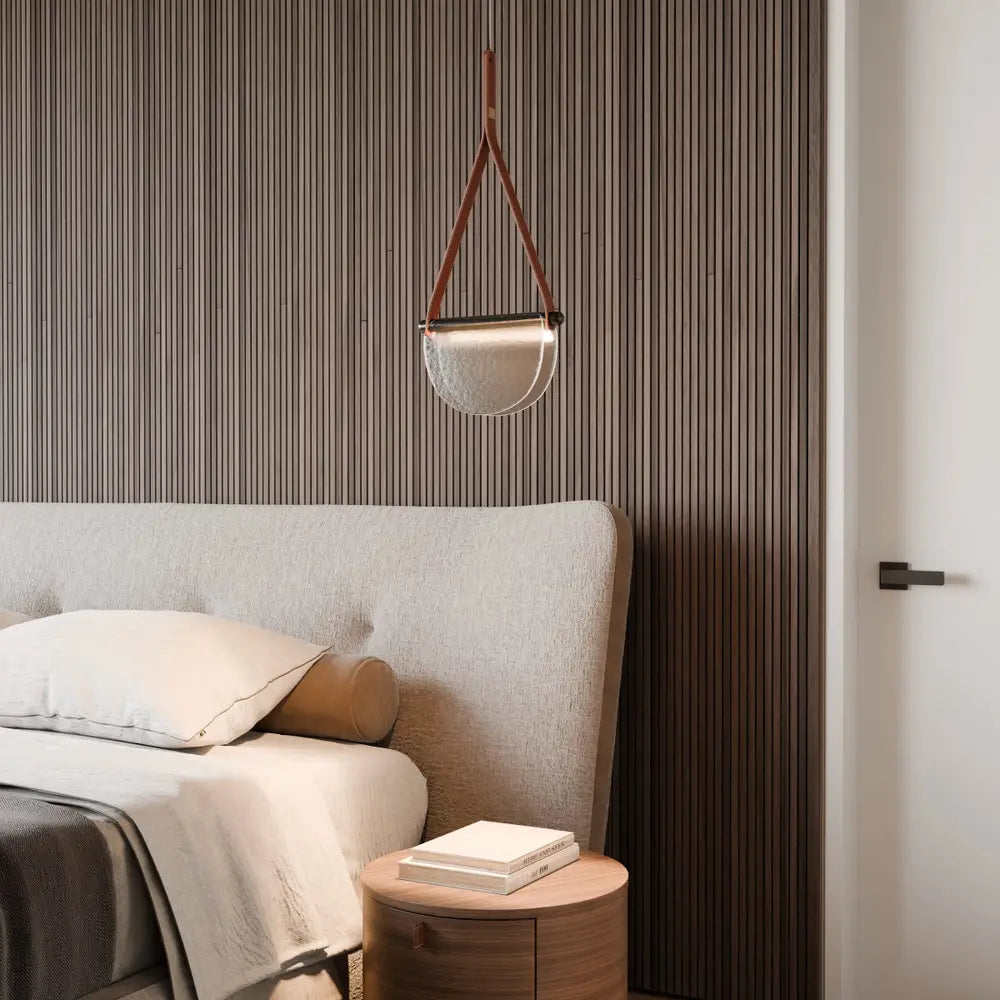 Clérachic pendant with a tan leather strap and half-round frosted glass shade hanging beside a bed against fluted walnut panelling.