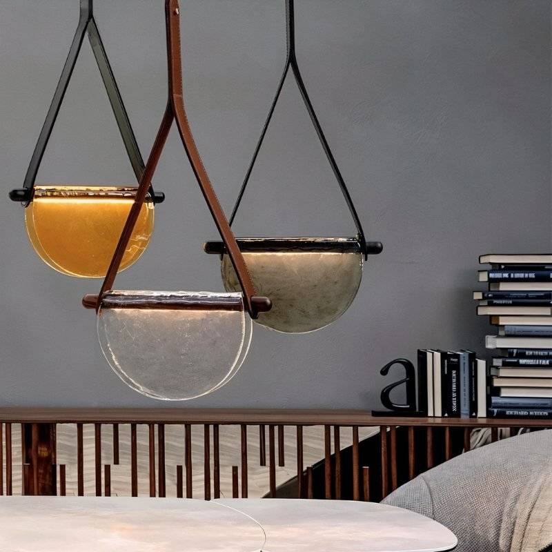 Three Clérachic leather-strap pendants in amber, clear and smoked glass hanging at different heights above a table by a bookshelf.
