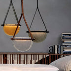 Three Clérachic leather-strap pendants in amber, clear and smoked glass hanging at different heights above a table by a bookshelf.