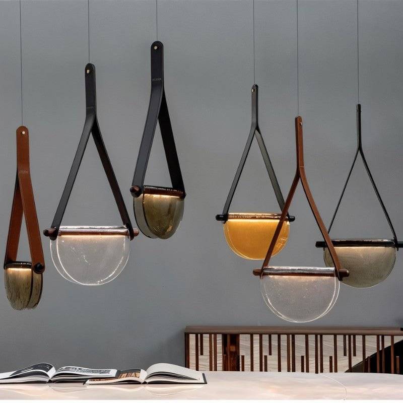 Group of Clérachic leather-strap pendants in smoked, clear and amber glass hung at varied heights over a marble table and open books.