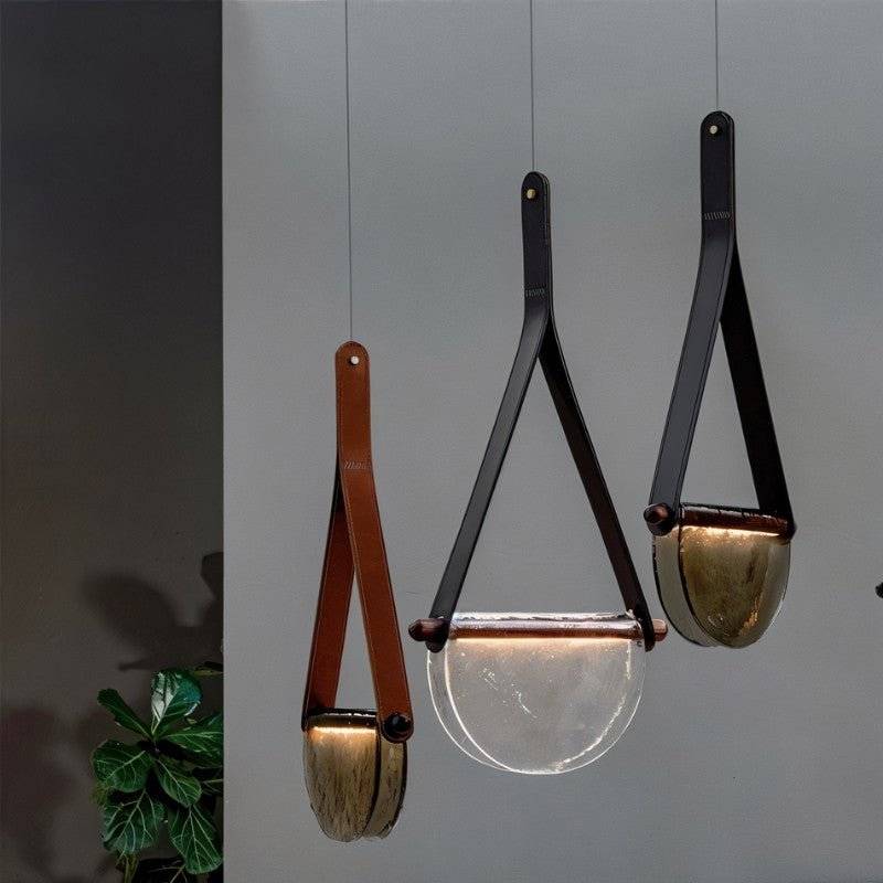 Three Clérachic pendants, two with black leather straps and one tan, holding half-round smoked and clear glass shades on a grey wall.