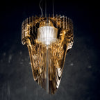 A Clérachic gold acrylic fringe pendant, lit from inside, seen against a dark backdrop with the stacked fins catching the light.