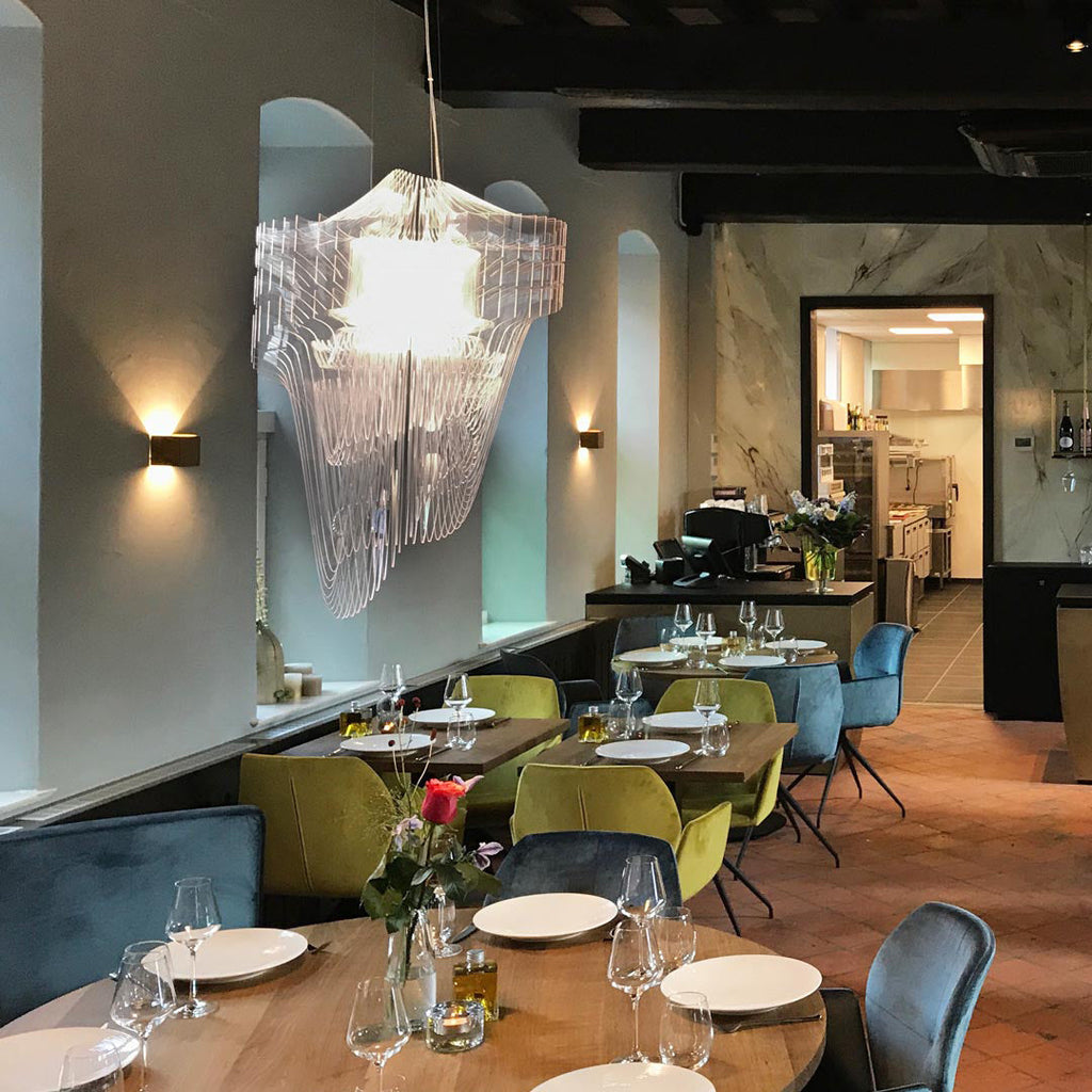 A clear acrylic fringe pendant from Clérachic hangs in a restaurant dining room above set tables and teal chairs, with wall lights behind.