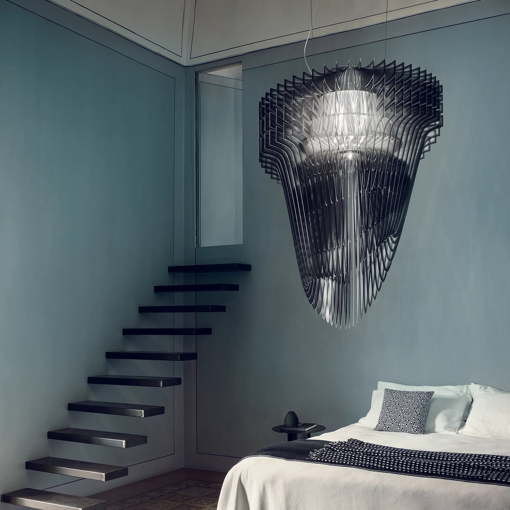 A Clérachic fringe pendant in black acrylic hangs in a tall blue-grey room beside a floating black staircase and a made bed.