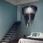 A Clérachic fringe pendant in black acrylic hangs in a tall blue-grey room beside a floating black staircase and a made bed.