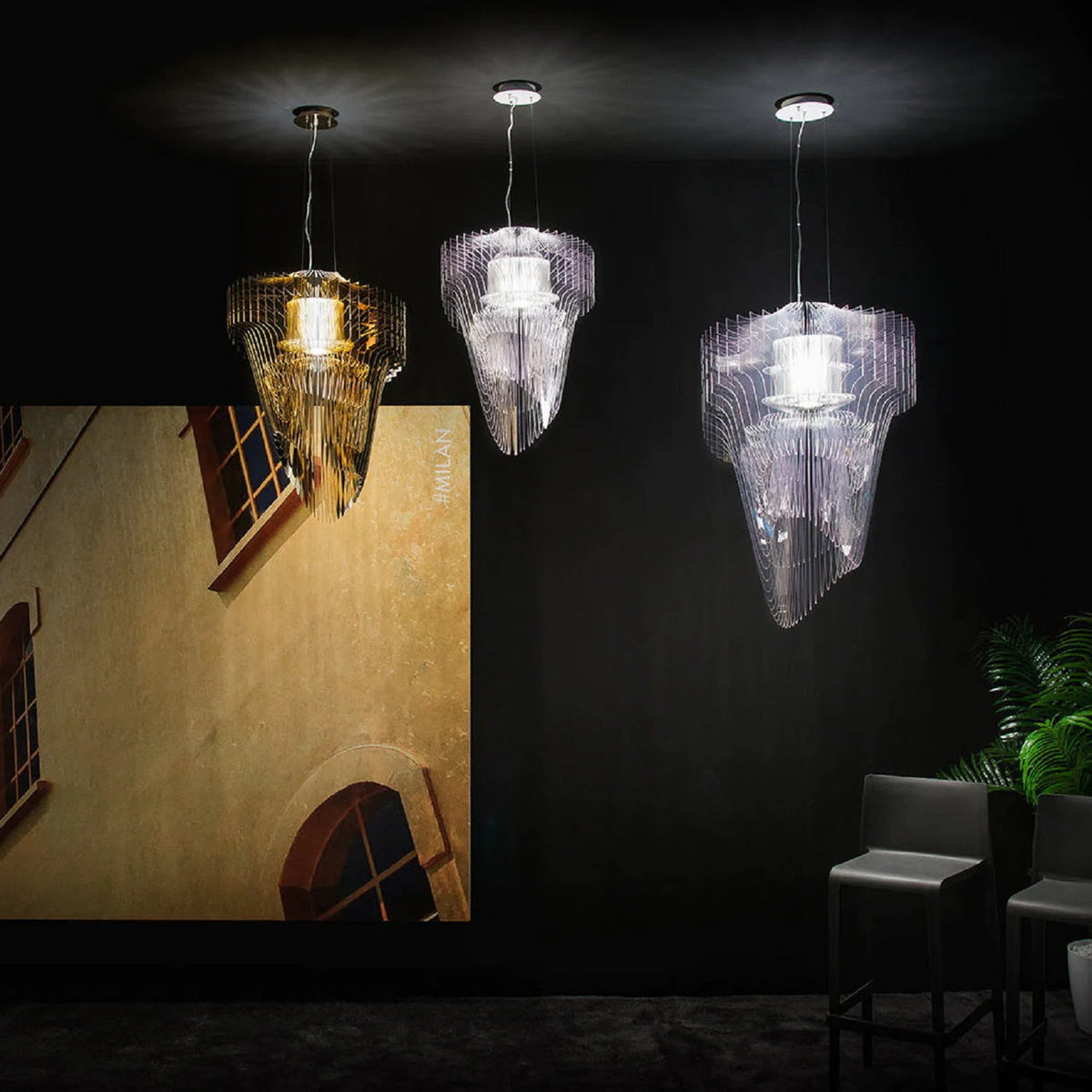 A gold pendant between two clear ones from Clérachic, all lit, hang in a black room in front of a warm yellow wall mural.