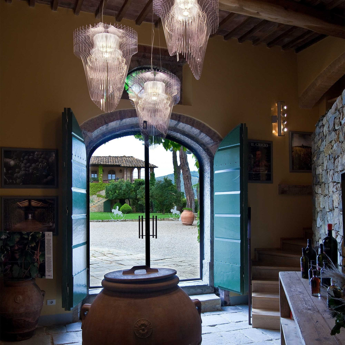 Several clear fringe pendants from Clérachic hang in a rustic hall with timber beams, in front of open green doors and a courtyard.