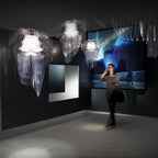 Three fringe pendants from Clérachic, mostly clear with one pale, hang in a dark gallery as a visitor passes in front of a large screen.