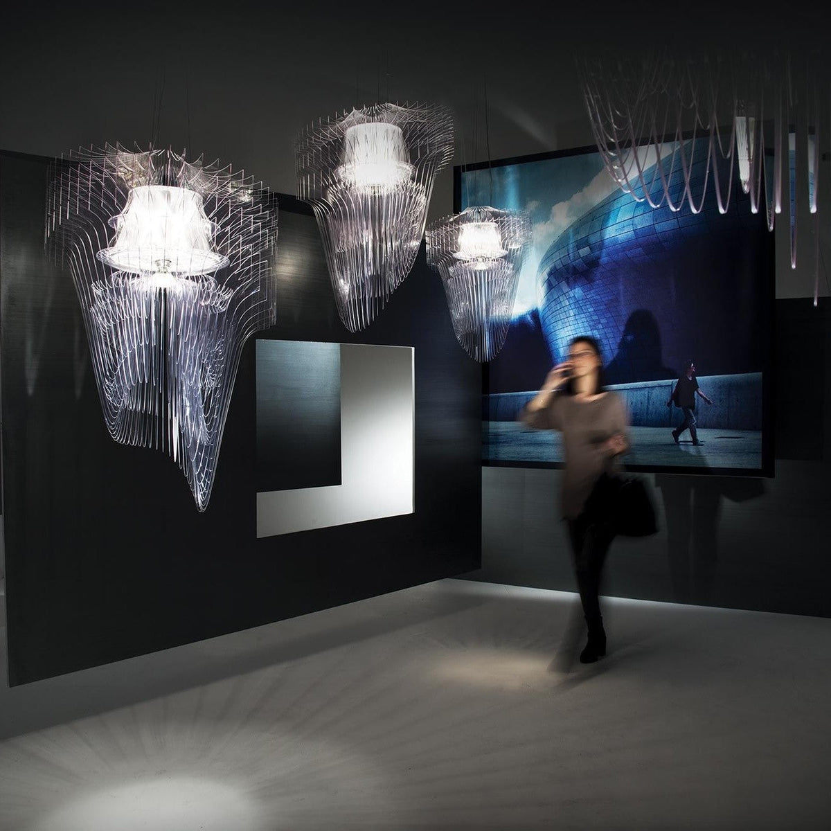 Three fringe pendants from Clérachic, mostly clear with one pale, hang in a dark gallery as a visitor passes in front of a large screen.