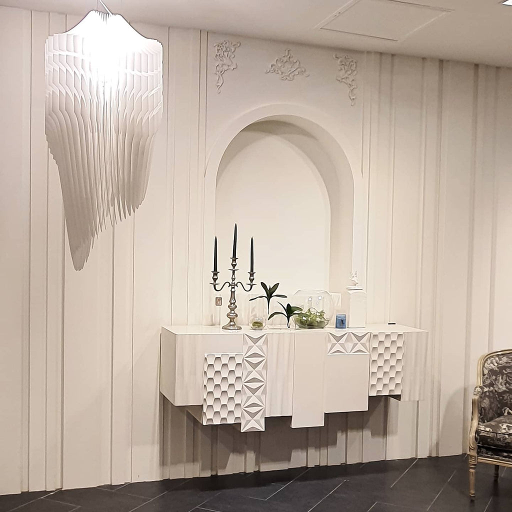 A pale, sculpted fringe light from Clérachic hangs at the top of the frame in a white room with an arched niche and a carved white sideboard.