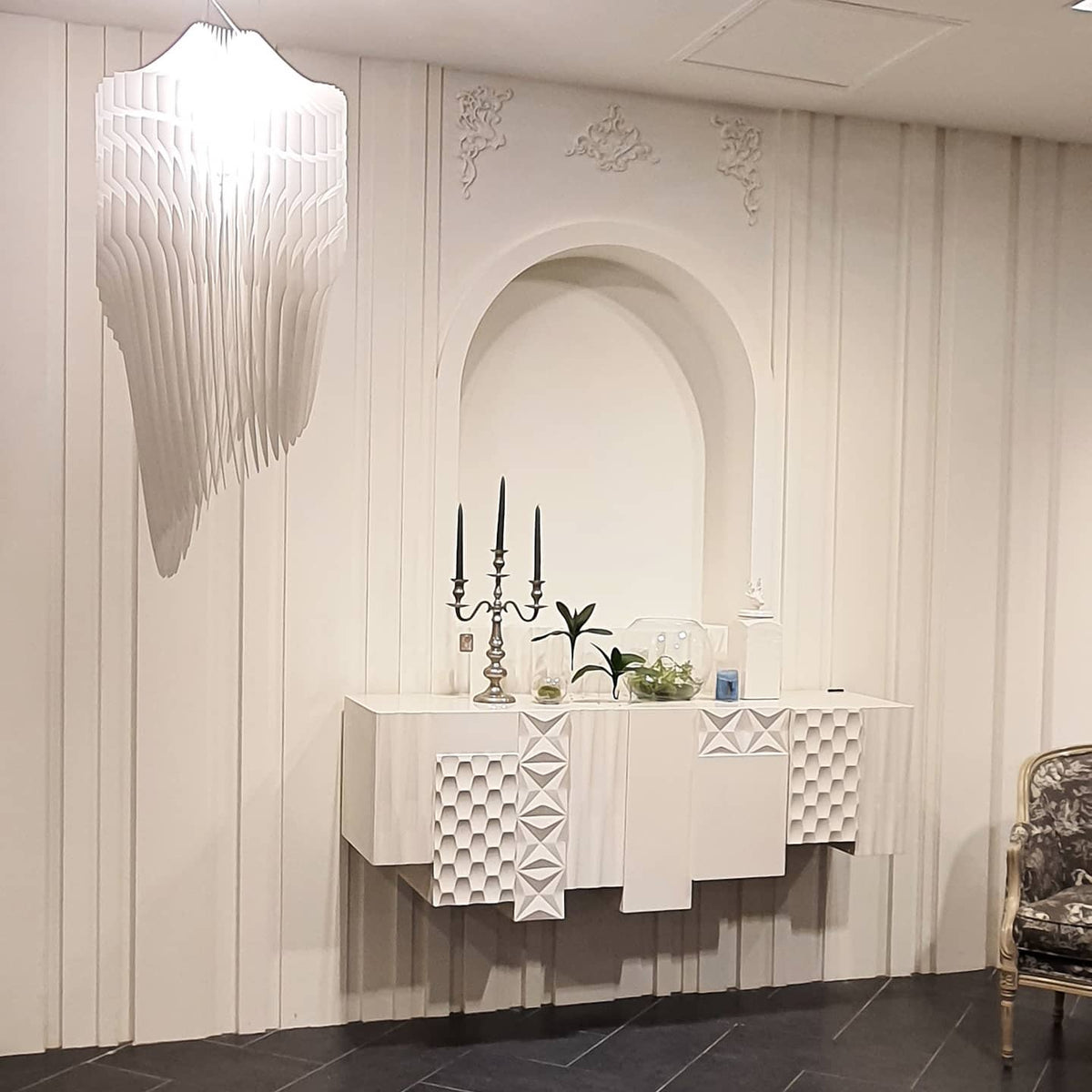 A pale, sculpted fringe light from Clérachic hangs at the top of the frame in a white room with an arched niche and a carved white sideboard.